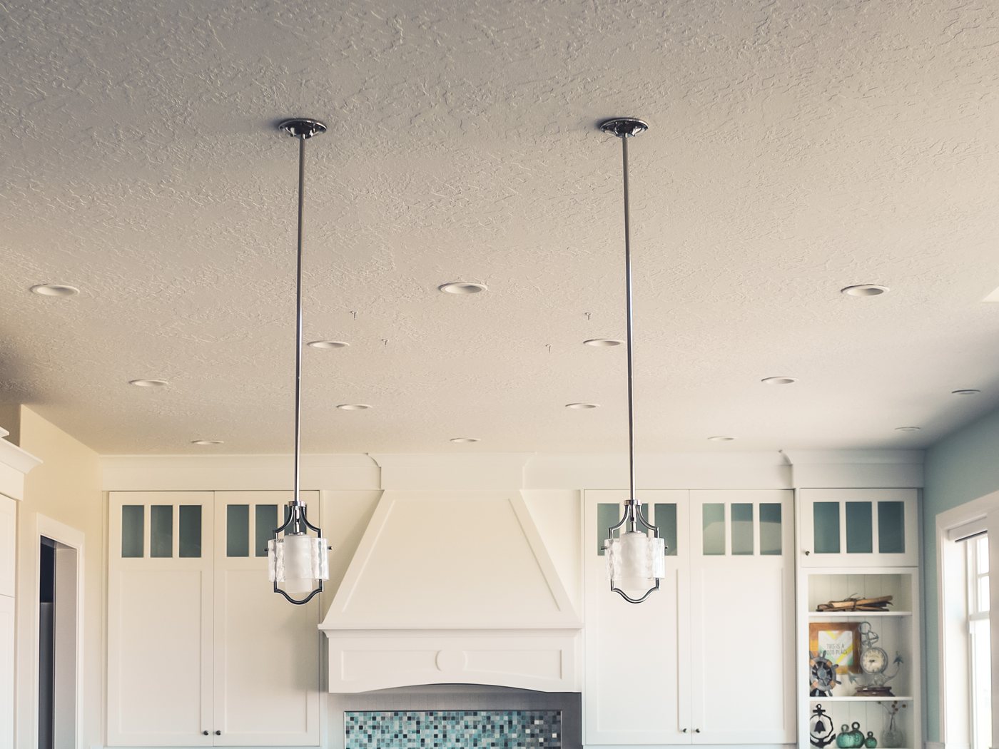 A kitchen ceiling showing recessed lighting and two pendant lights over an island, with cabinetry below.
